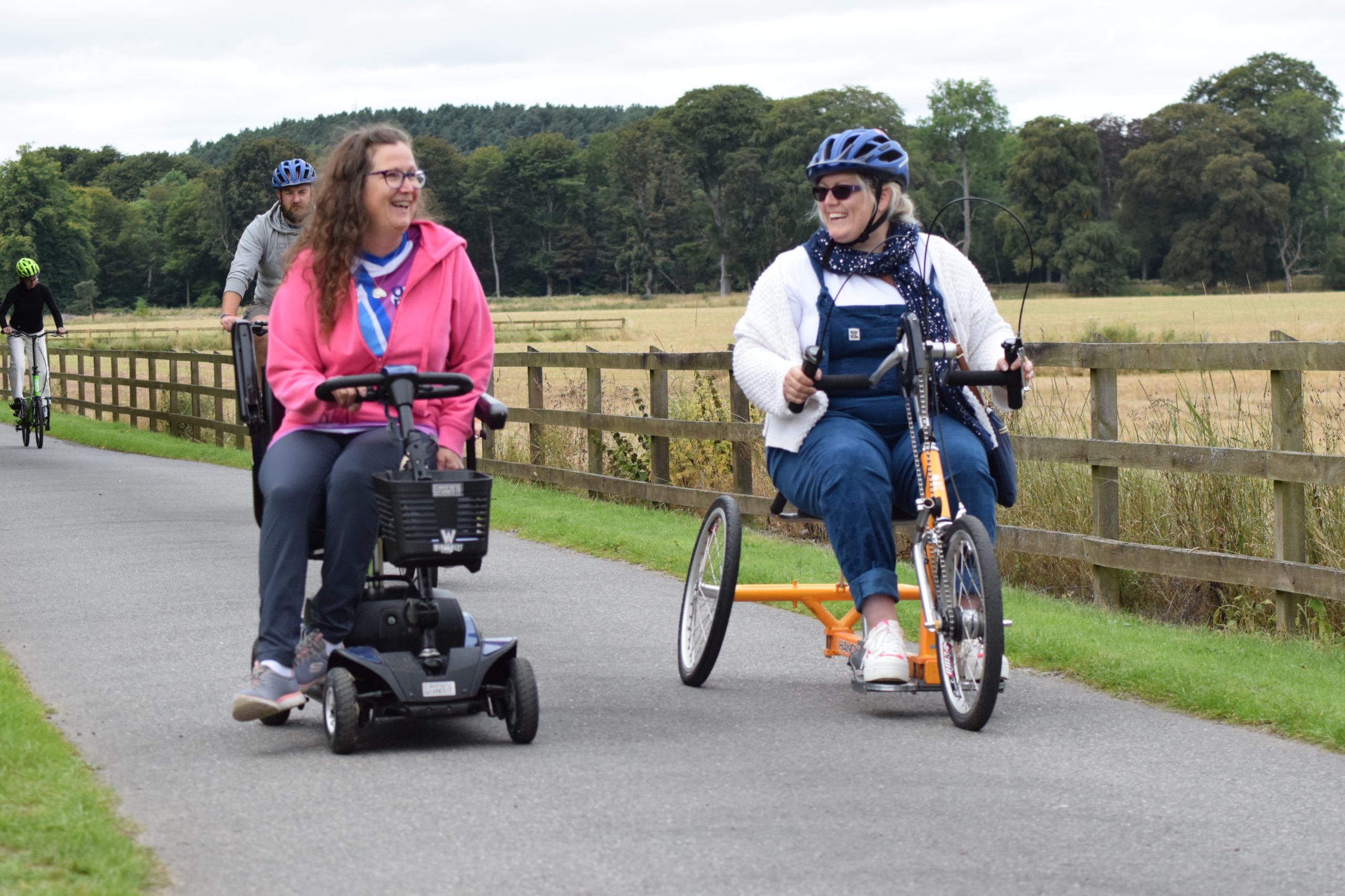 People using various types of transport options on a shared use path, front and centre a person on a mobility scooter and a person using a hand cycle